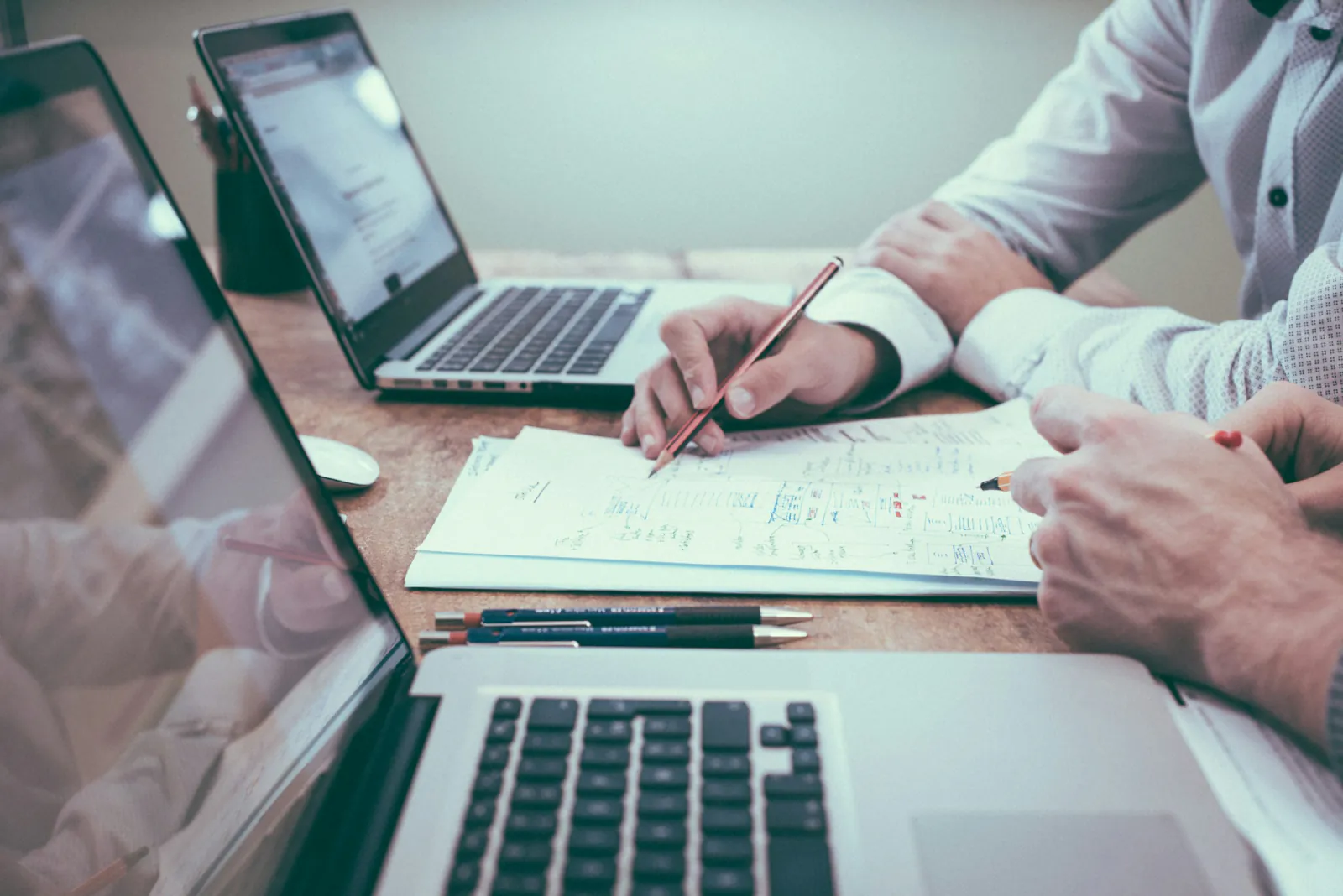 Two people working through handwritten notes and printed papers beside open laptops on a wooden desk