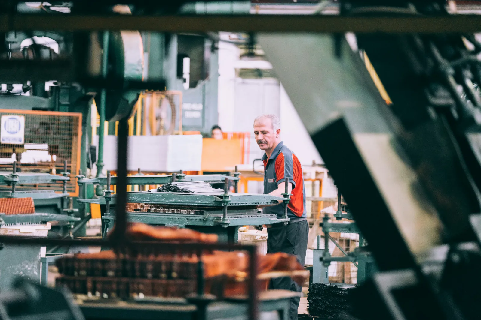 Machine operator in a grey and red work shirt tending industrial equipment on a busy factory floor