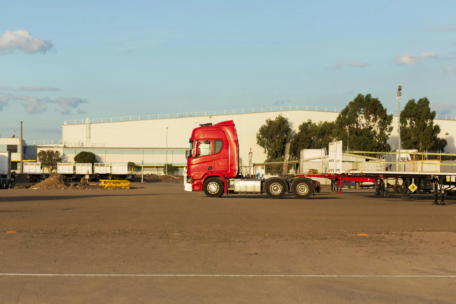Red prime mover and empty trailer parked in a gravel yard beside a large warehouse in warm late afternoon light, Altona, Victoria
