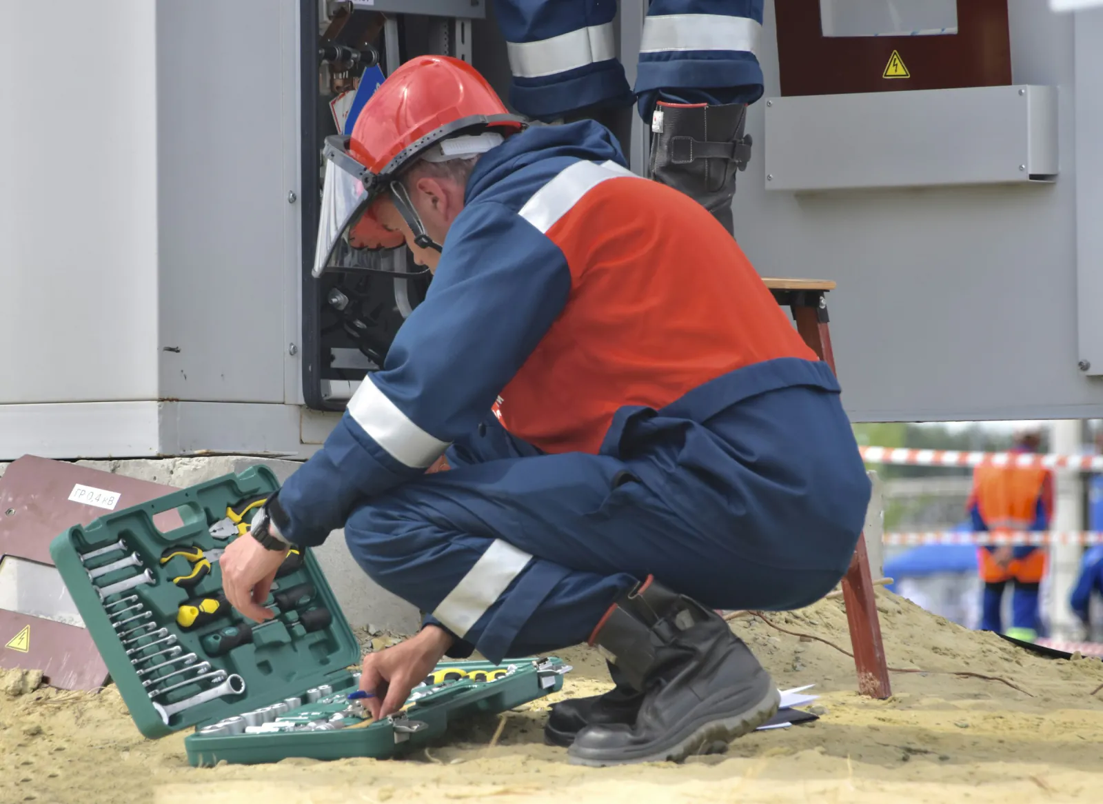 Service technician in a hard hat and high-visibility coveralls selecting tools from a kit beside an open electrical cabinet