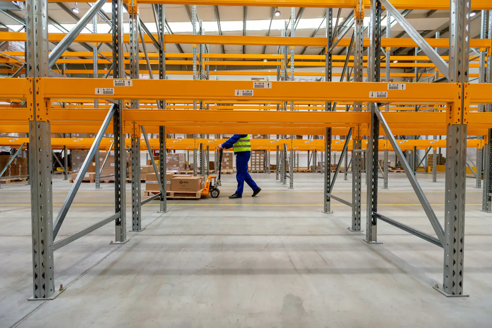 Warehouse worker moving a pallet with a hand pallet jack between rows of orange pallet racking